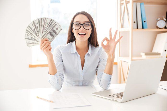 business-woman-dressed-in-white-shirt-sitting-in-her-office-and-holding-money-in-hand-while-making-okay-gesture 11 Ways You Can Grab Extra Money This Week